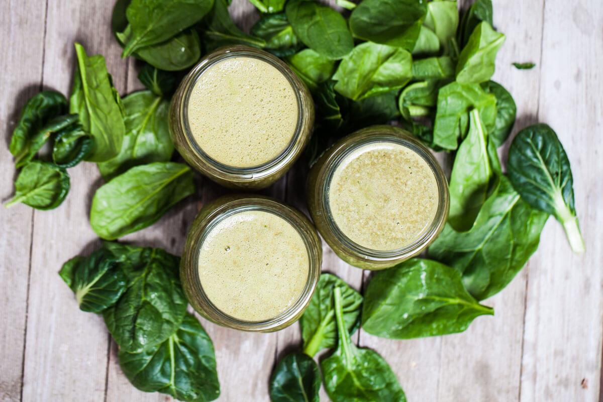 Three green smoothies in mason jars surrounded by spinach leaves.